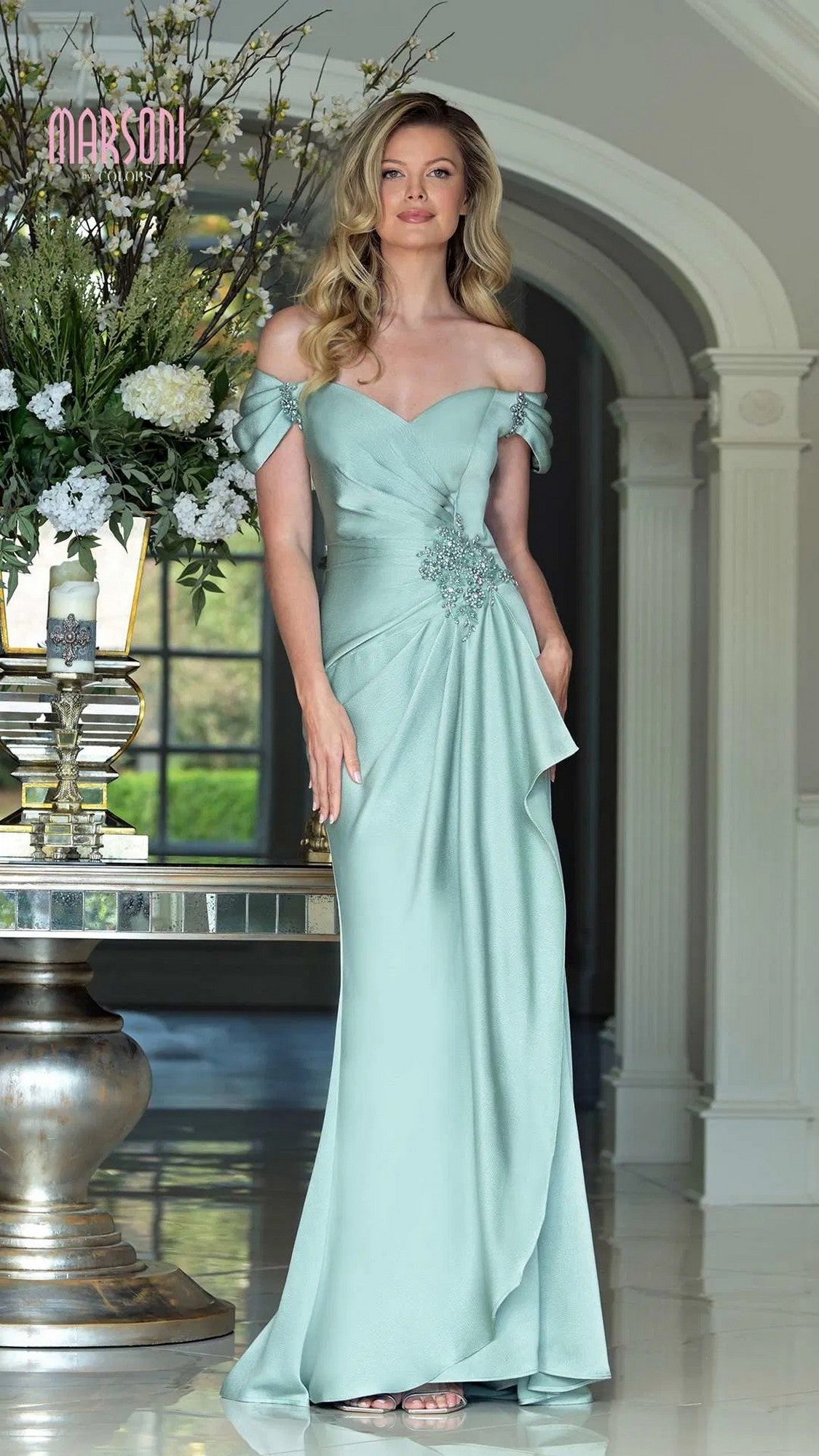 Wearing the Marsoni MV1351 Long Formal Dress in light blue with floral embellishments, a woman stands indoors by a decorative table and large floral arrangement, her elegant off-the-shoulder chiffon gown enhancing the scene.
